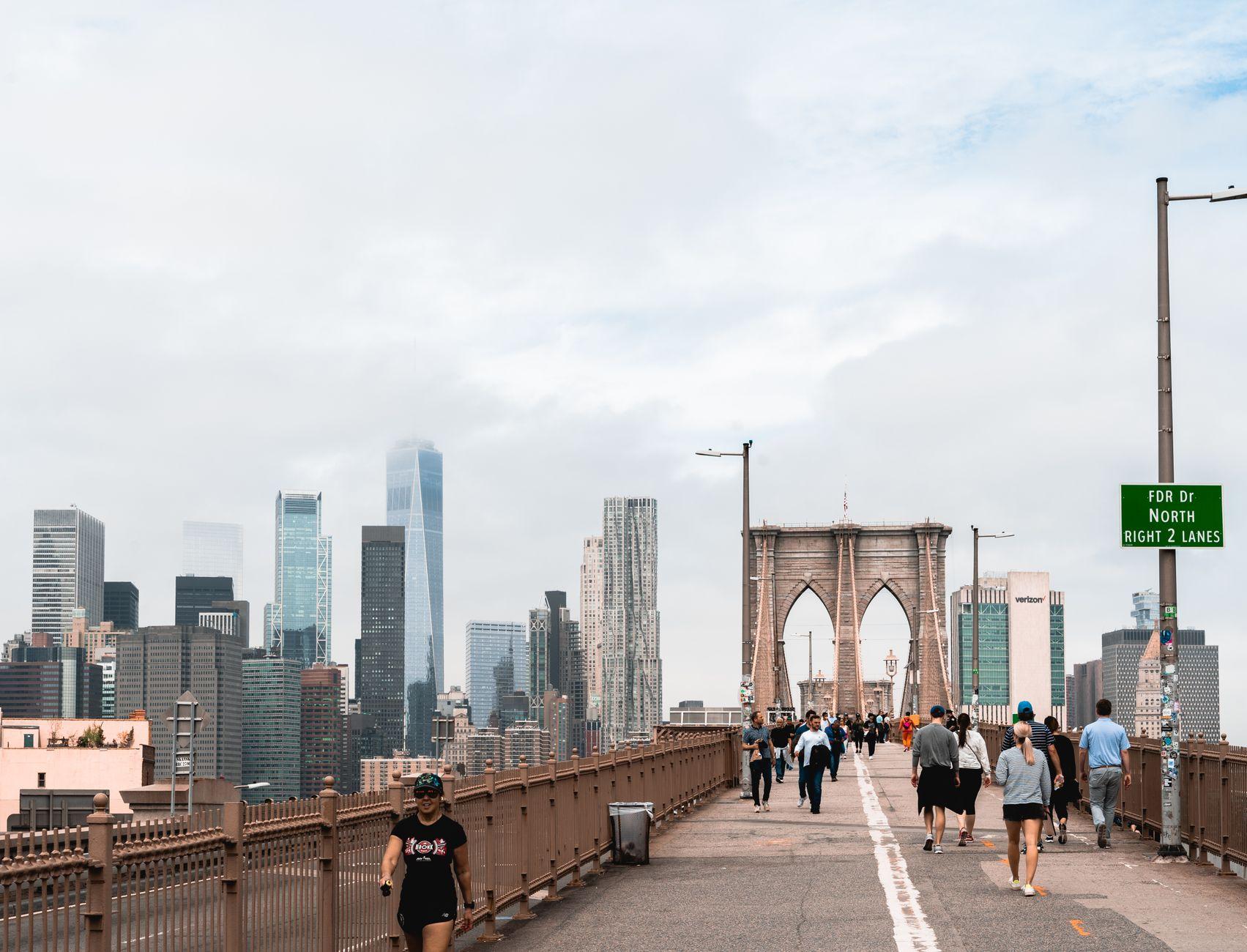 People walking on Brooklyn Bridge with Manhattan skyscrapers in the background under cloudy skies.
