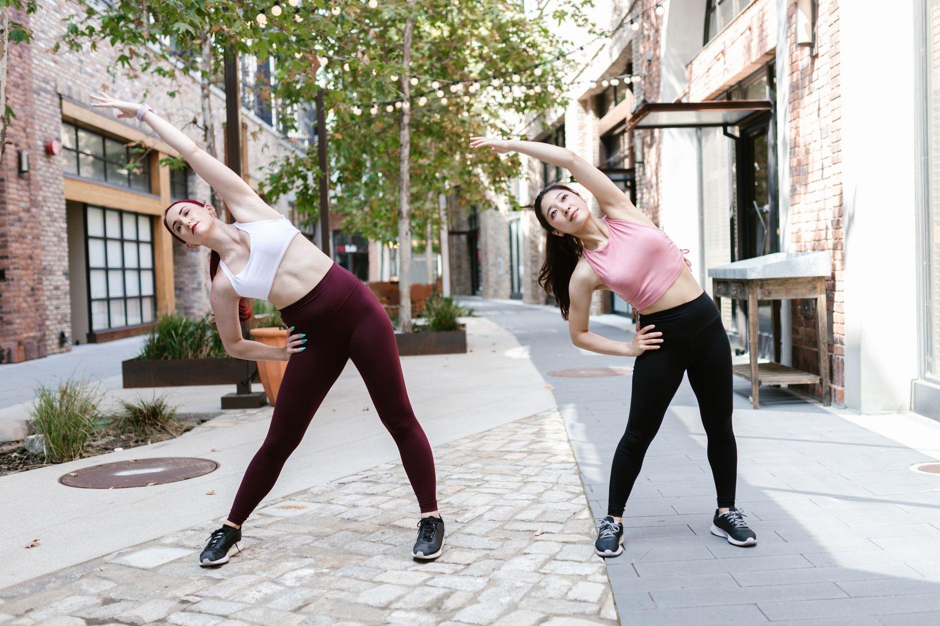 Two women perform stretching exercises outdoors in an urban setting, promoting a healthy lifestyle.