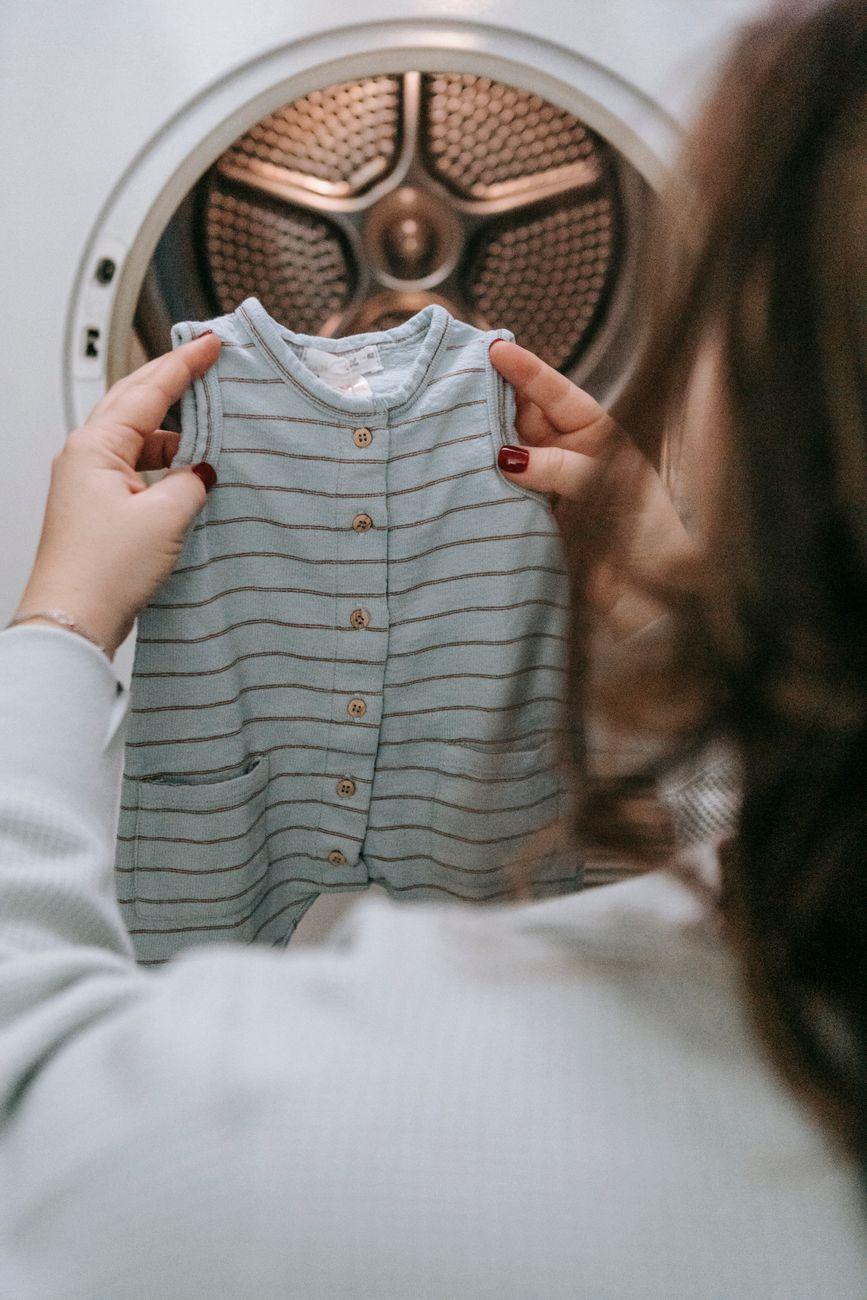 Back view of unrecognizable female with baby attire in hands near modern washing machine during laundry routine in light room