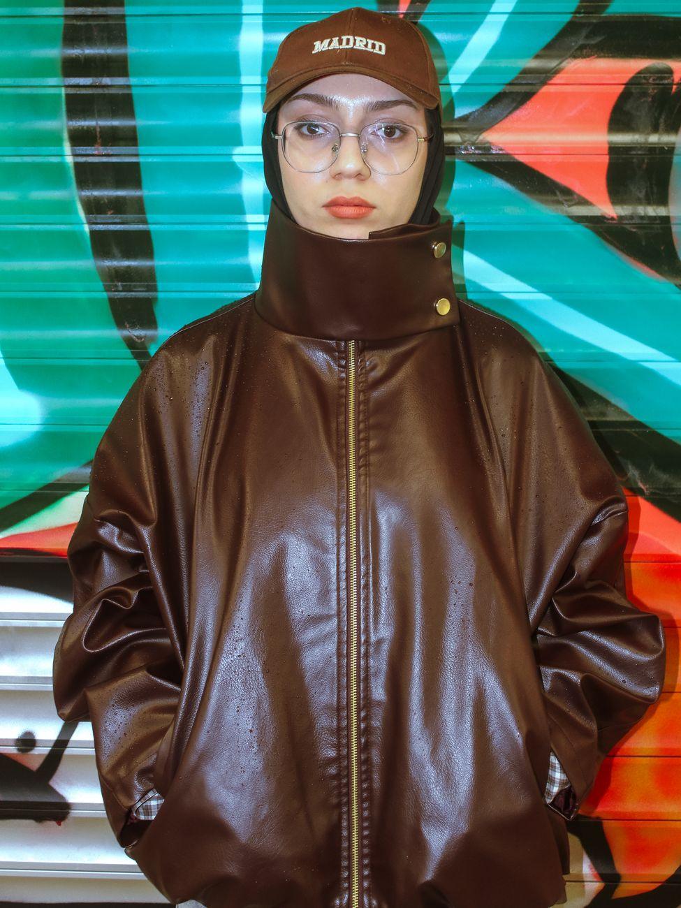 Fashionable woman in a dark brown leather jacket and cap posing against graffiti backdrop.